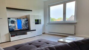 Ett modernt vardagsrum med en platt-TV, vita skåp, en grå soffa och ett fönster med utsikt över hus.