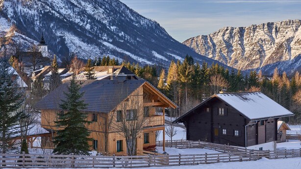 Eine Holzhütte mit schneebedecktem Dach, umgeben von einem Holzzaun, vor einer Kulisse aus schneebedeckten Bergen und blauem Himmel.