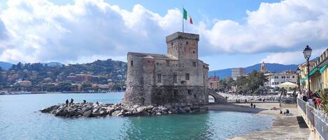 A coastal town with a historic stone tower, clear blue waters, and a paved promenade with buildings and palm trees.