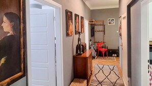 Un couloir avec une porte blanche, un meuble en bois et une chaise rouge. Des tableaux encadrés ornent le mur, et un luminaire est fixé au plafond.