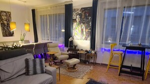 Un salon moderne avec un canapé gris, un fauteuil beige, une petite table basse et une chaise jaune. Deux tableaux ornent les murs, ainsi qu’une plante en pot et une fenêtre avec des rideaux.