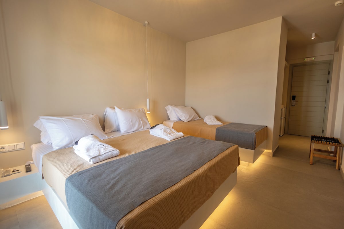 A hotel room with two beds, a bedside table, a lamp, and a small wooden stool.