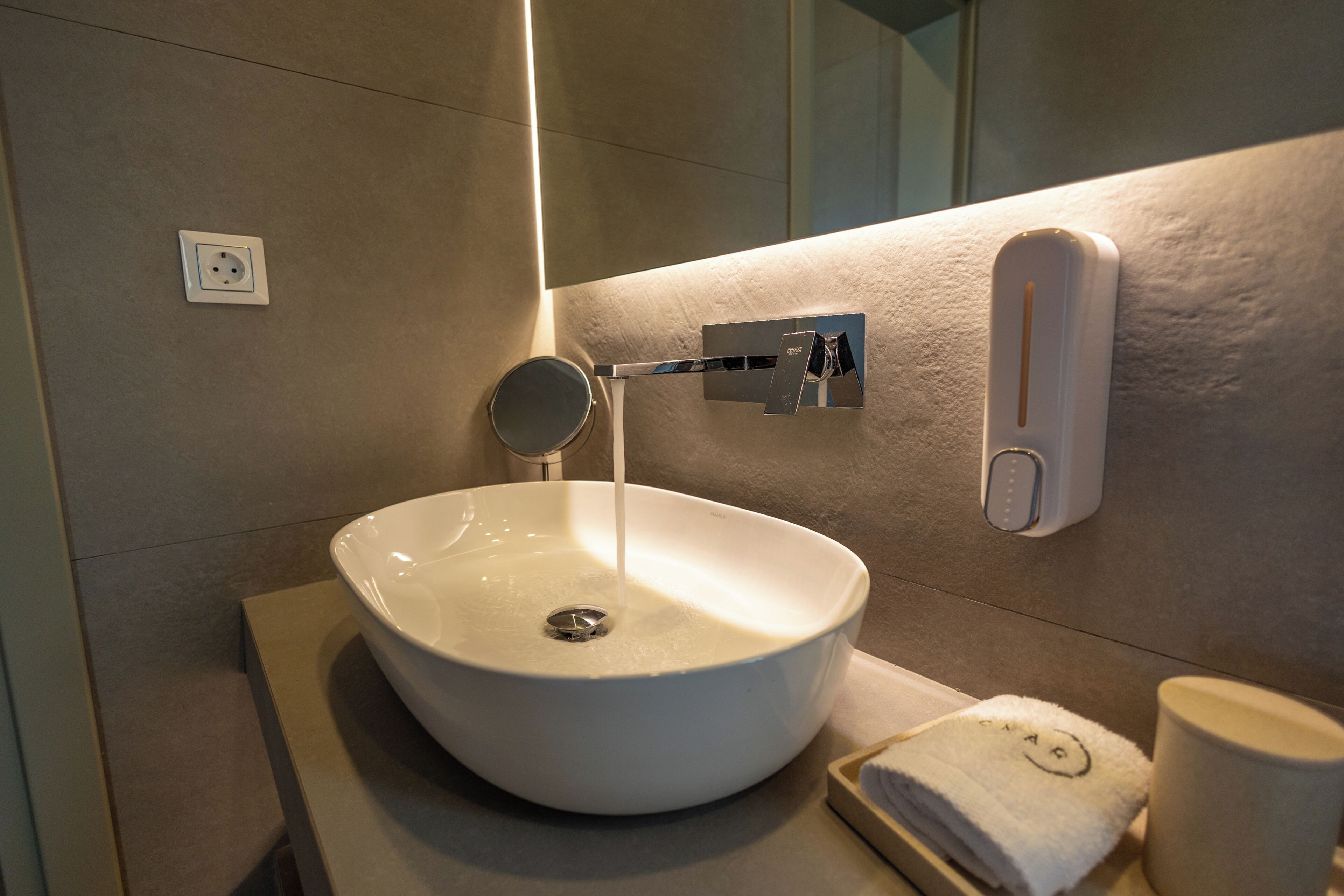 Un baño con lavamanos blanco, espejo, toalla y dispensador de jabón.