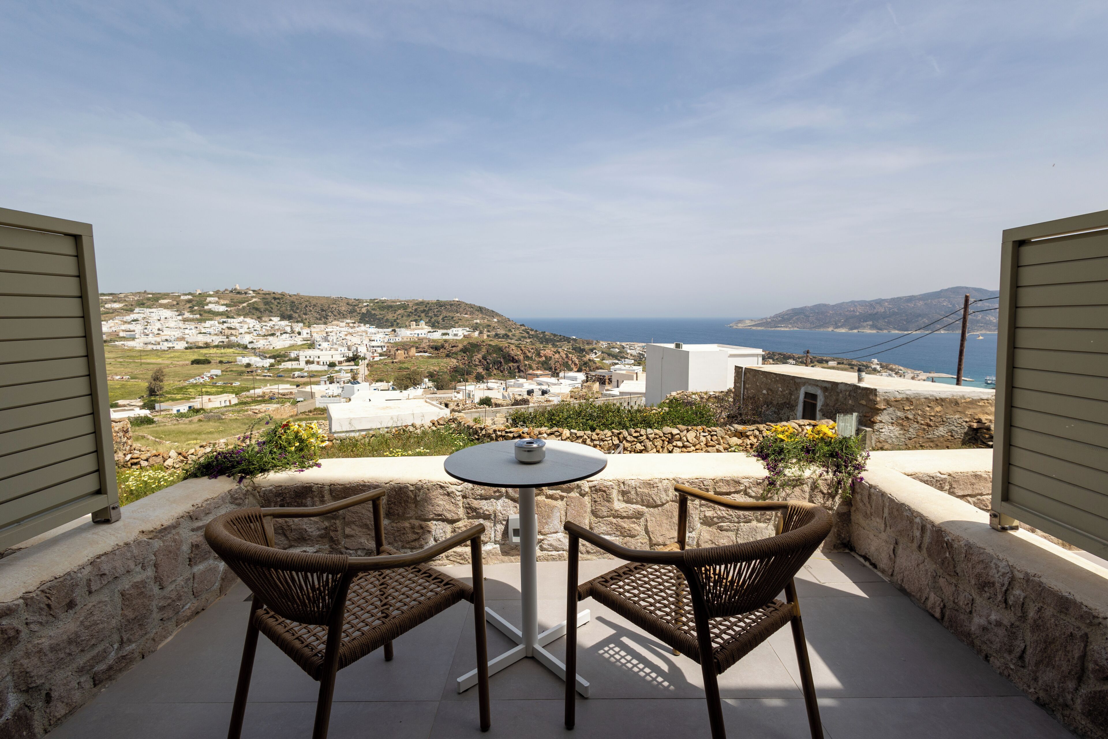 A balcony with wicker chairs and a table, overlooking a coastal village with white buildings and a view of the sea.