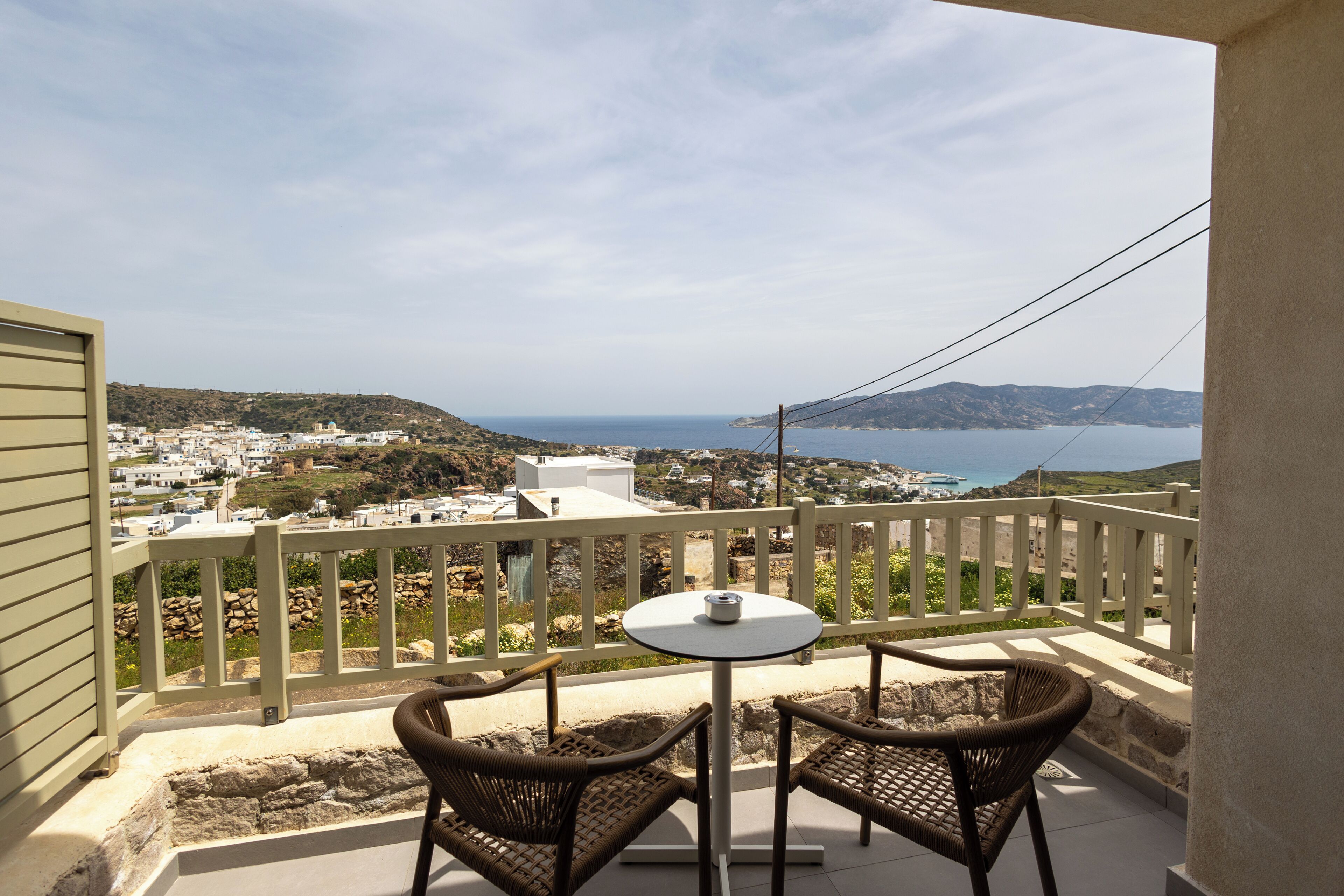 A balcony with wicker chairs and a small table, overlooking a coastal town and sea.