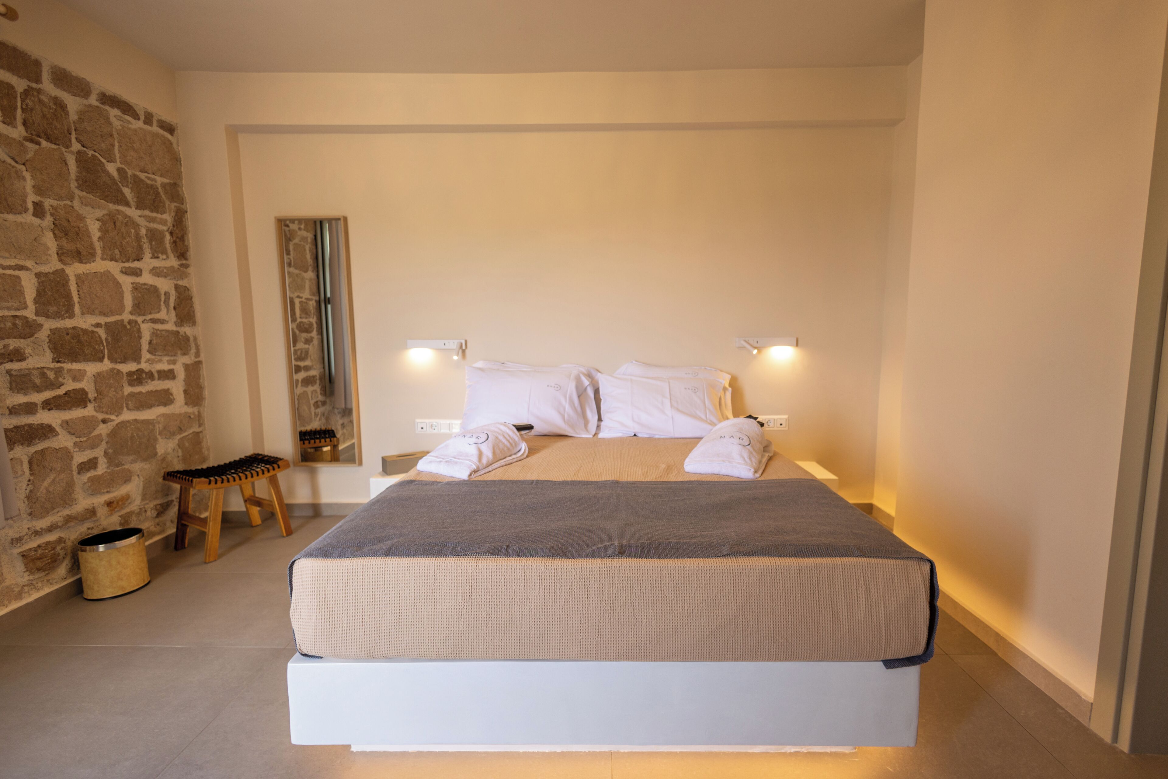 A bedroom with a bed, bedside lamps, a wooden stool, and a stone wall.