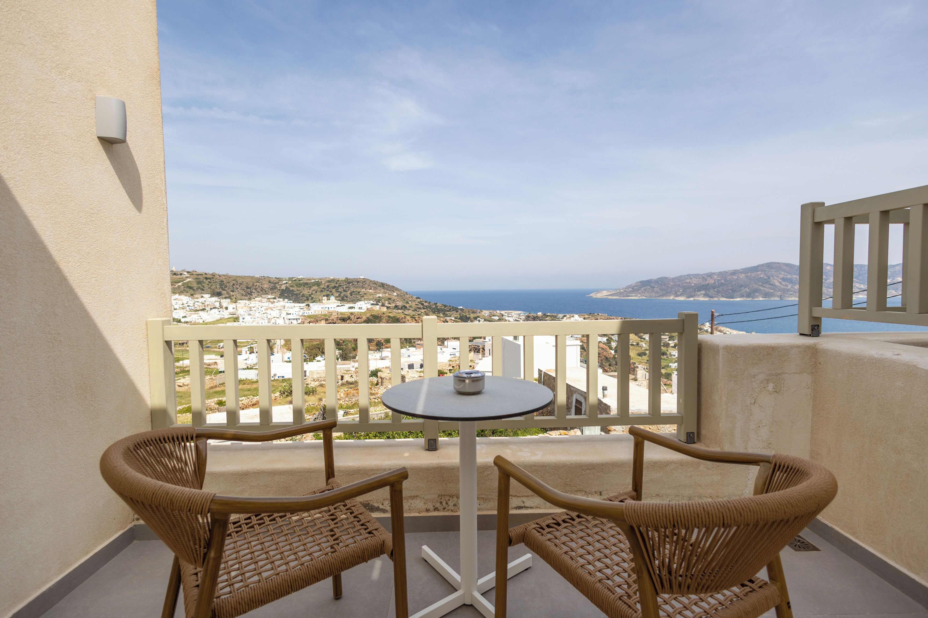 A balcony with wicker chairs and a small table, overlooking a coastal landscape with buildings and hills.