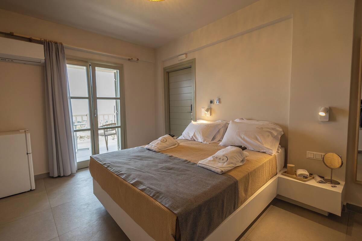 A hotel room with a bed, a small bedside table, a window with curtains, and a door.