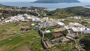 An aerial view of a coastal village with white buildings, a stone wall, and a view of the sea and distant island.