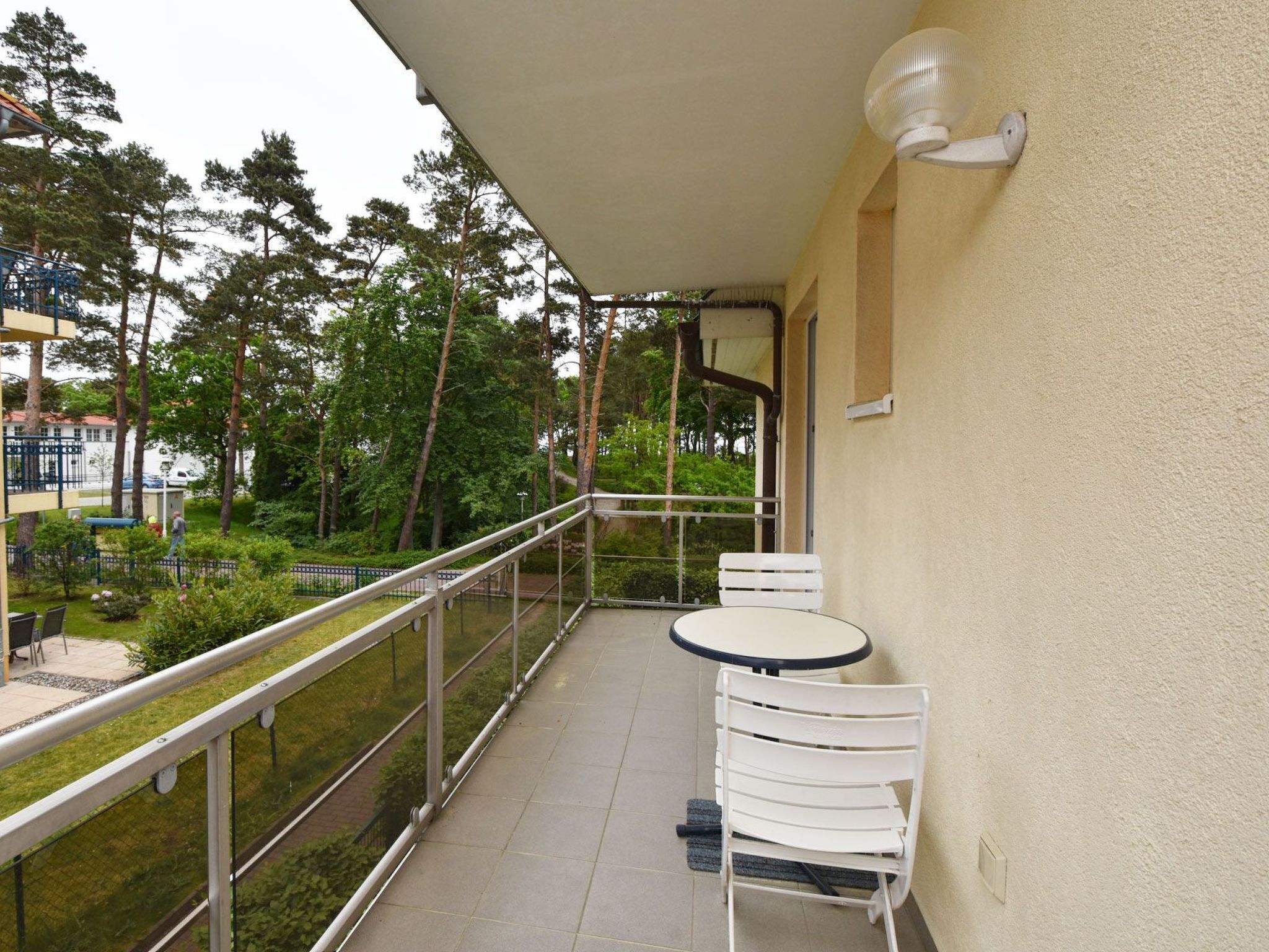 A balcony with a table and two chairs, a railing, and a view of a green area with trees.