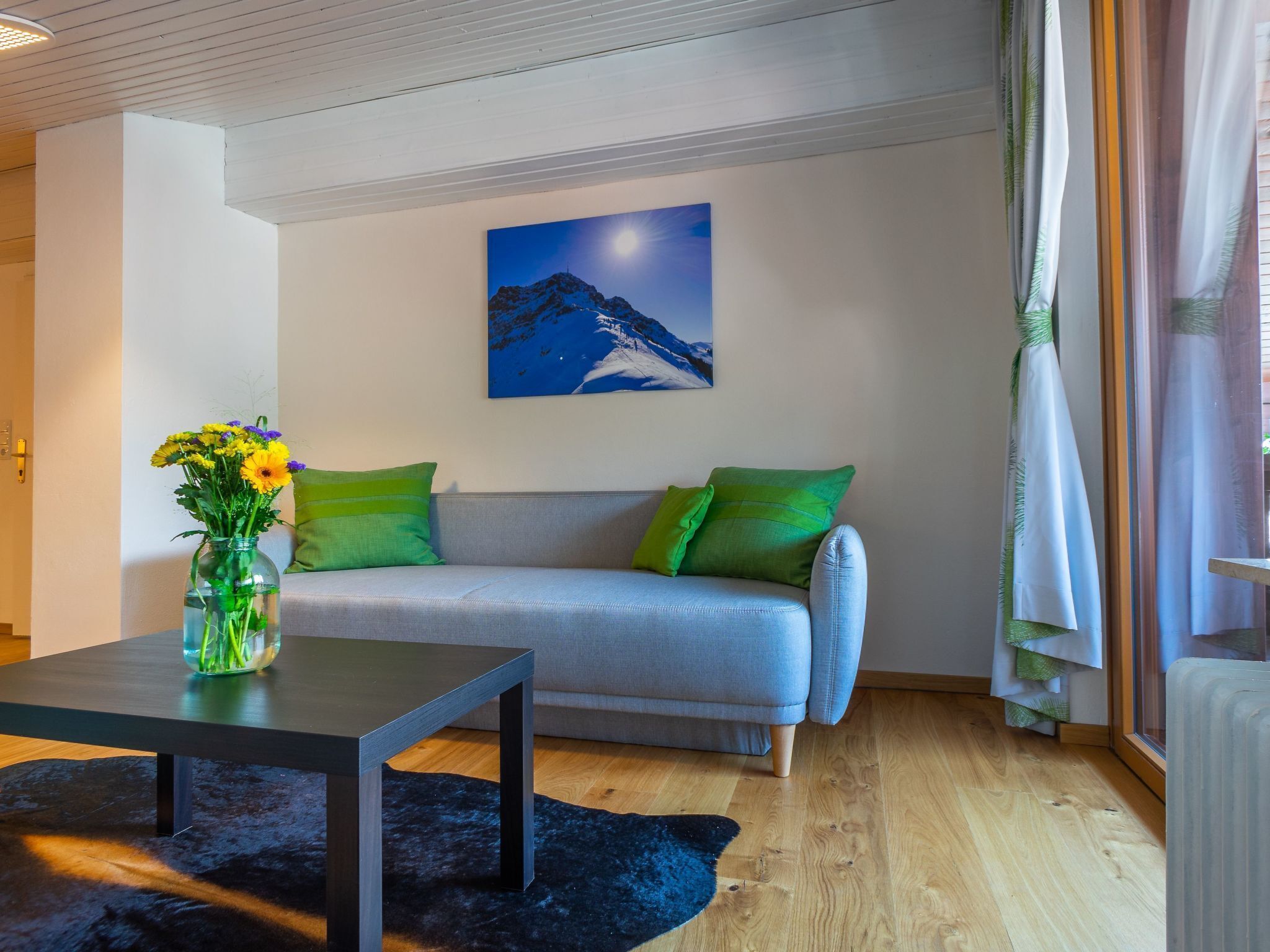 Un salon moderne avec un canapé gris, des coussins verts, une table basse noire et un vase rempli de fleurs.