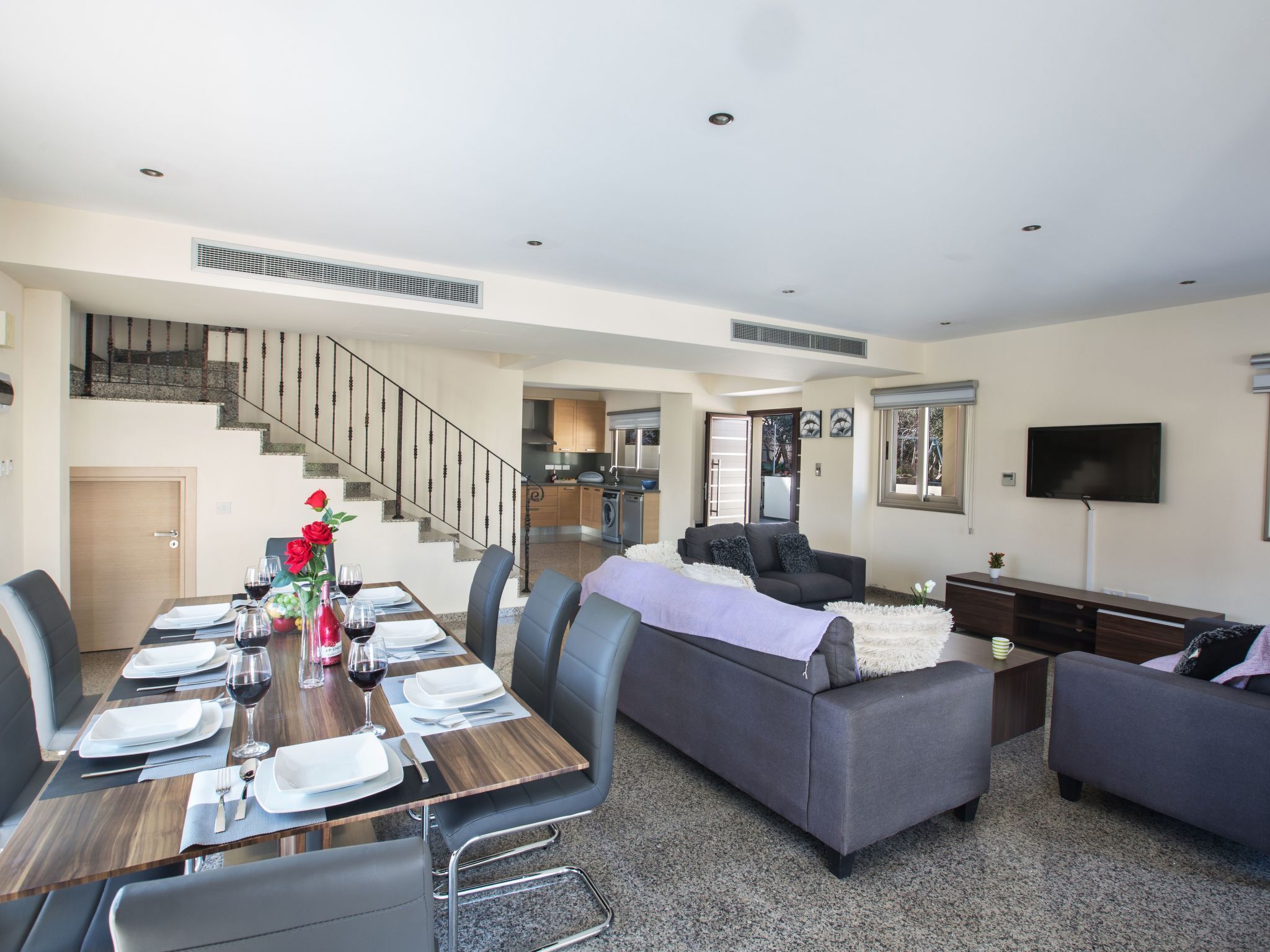 Un salon moderne avec un coin repas, des canapés gris, un escalier et une cuisine en arrière-plan.