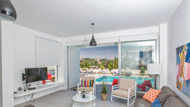 A modern living room with a TV, seating, a coffee table, and a view of a swimming pool and buildings. - Protaras Greco Villa CCV1 mit beheiztem Pool by Interhome (Paralimni)