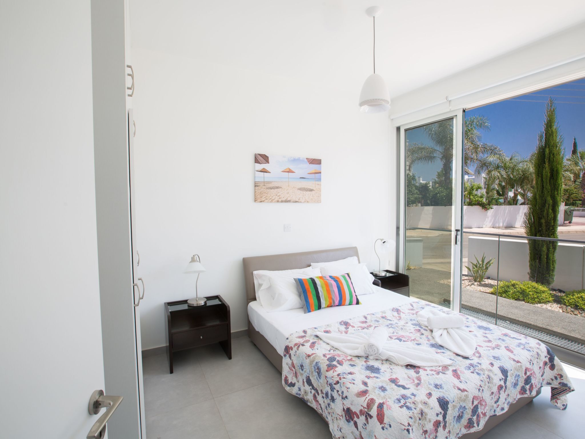 Une chambre avec un grand lit, une table de chevet, une lampe, un tableau représentant une plage et une vue sur l’extérieur.