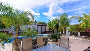 An outdoor patio with a glass table and chairs, surrounded by palm trees and a swimming pool. - Protaras Ferienvilla AV6 by Interhome (Protaras)