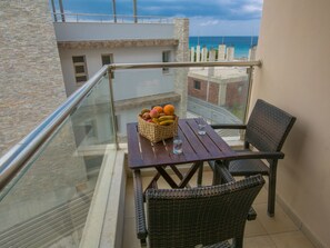 Un balcon avec des fauteuils en osier, une table sur laquelle se trouve un panier de fruits et des verres, et une vue sur la mer et les bâtiments.