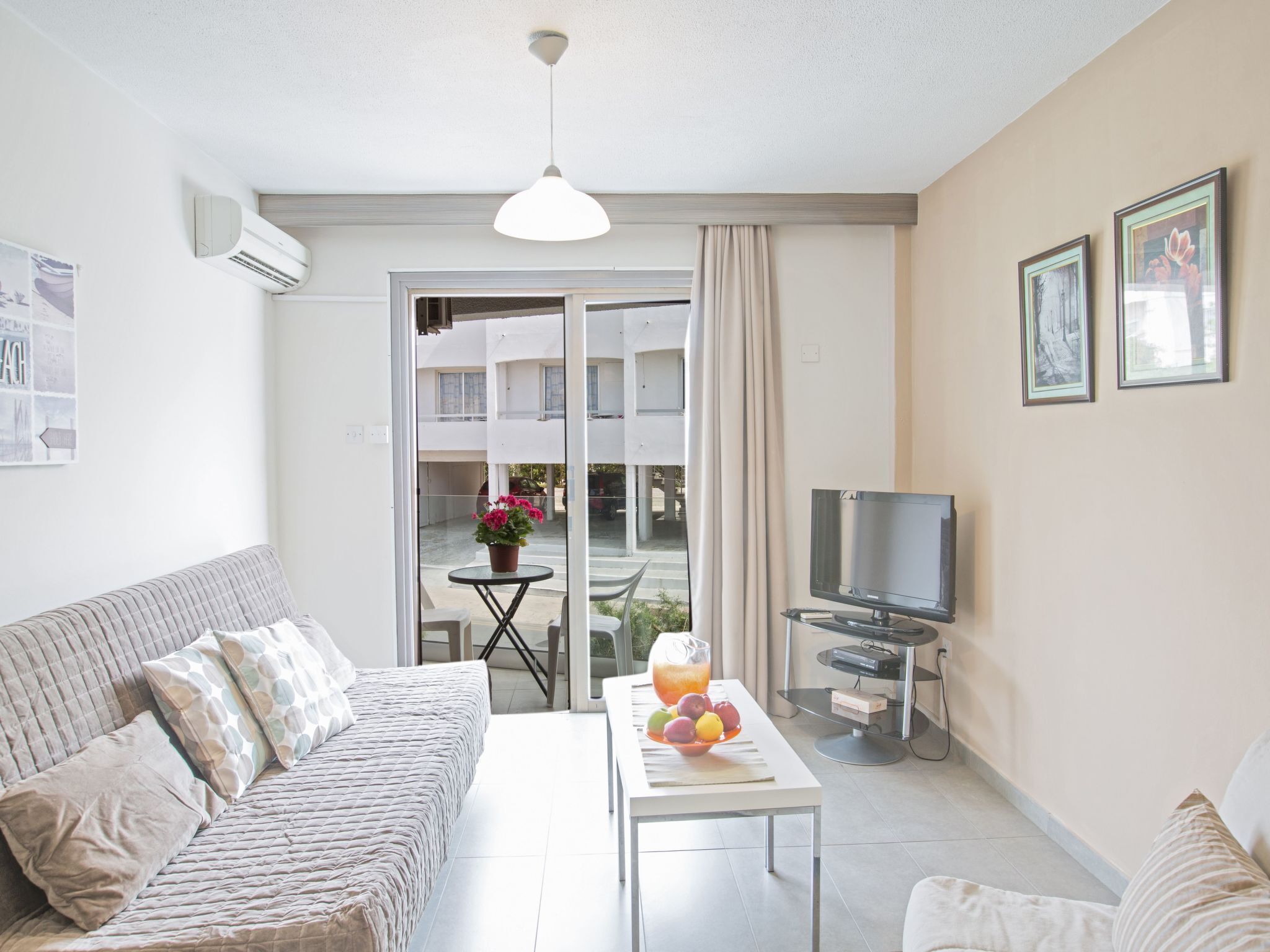 A modern living room with a sofa, a coffee table with fruit, a TV, and a balcony with a view of buildings.