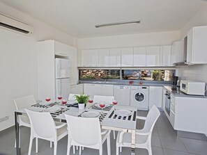 Cocina moderna con armarios blancos, horno empotrado, microondas y lavadora. Hay una mesa de comedor con sillas blancas y una planta en maceta.