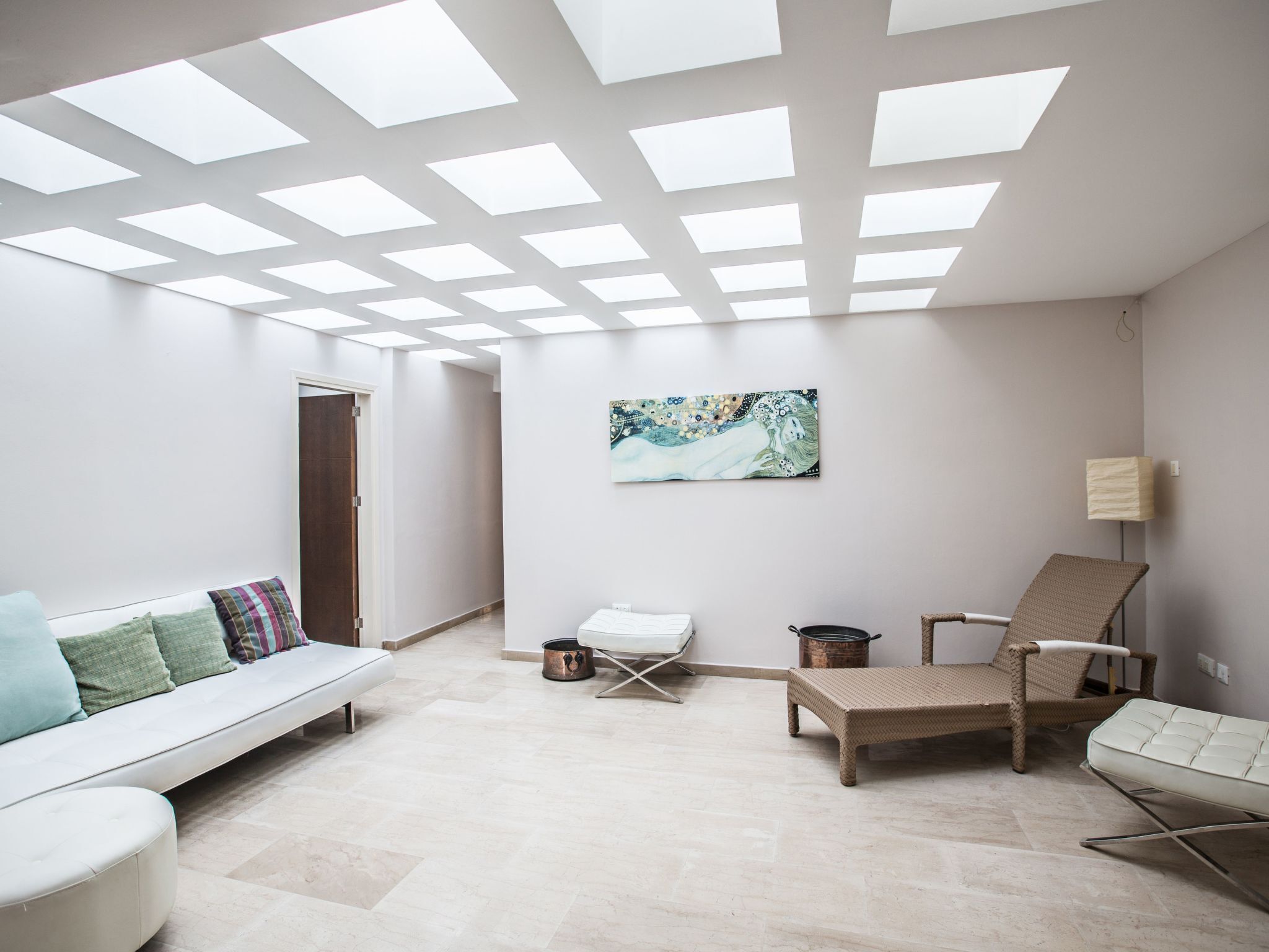 A spacious room with a high ceiling, skylights, a white sofa with cushions, a wicker chair, a small table, and a painting on the wall.