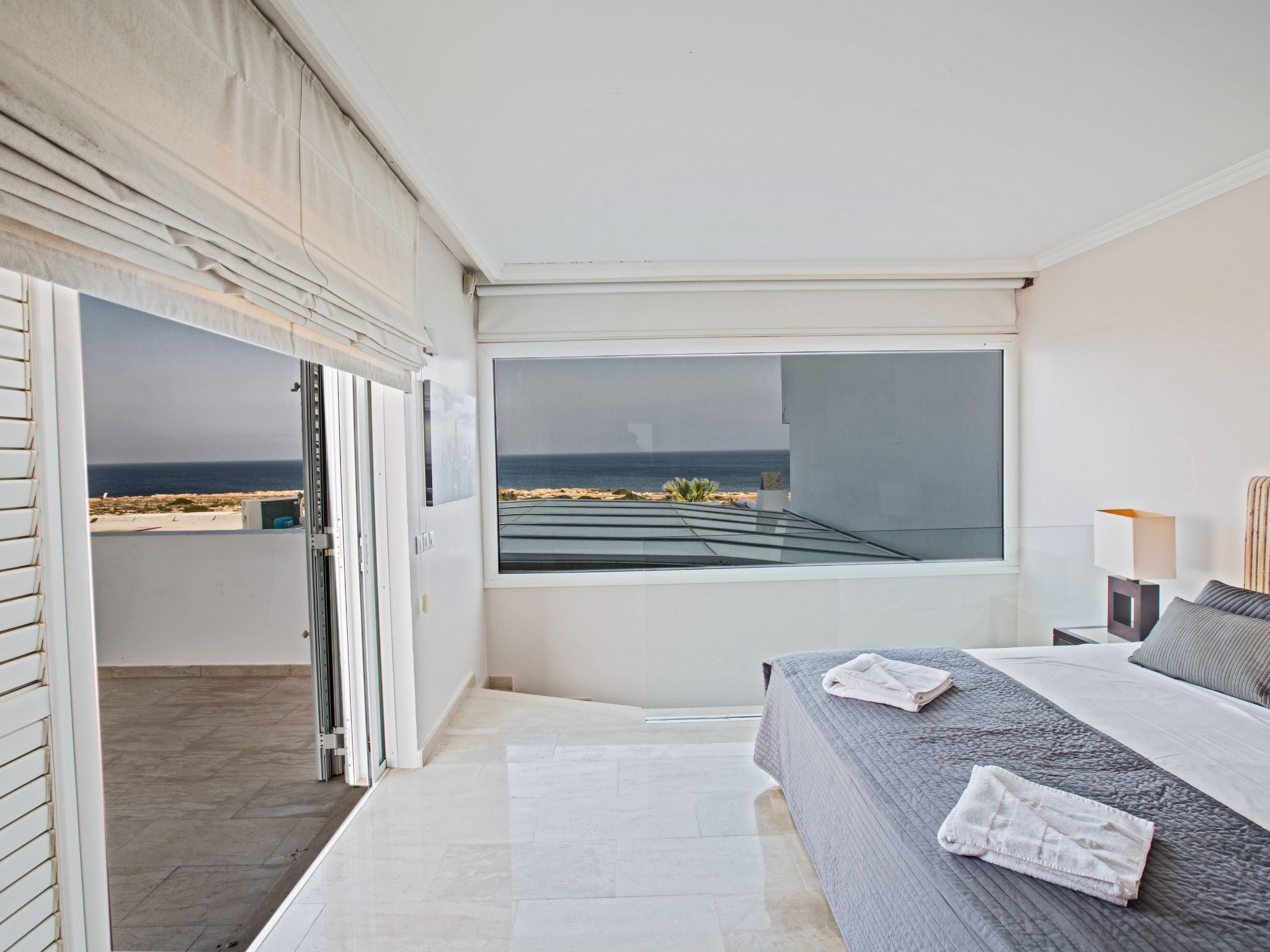 A modern bedroom with a large window overlooking a beach, a bed with a grey quilt, and a bedside table with a lamp.
