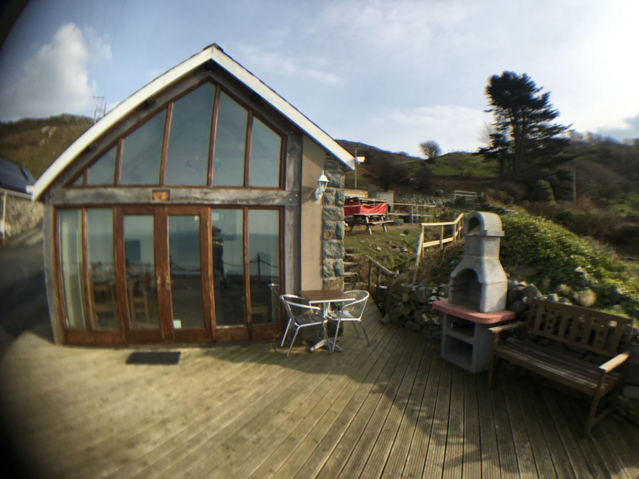 Eine Holzterrasse mit einem gläsernen Gebäude, einem Steinkamin und Gartenmöbeln.