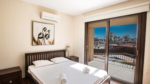 Ein Hotelzimmer mit Bett, Nachttisch, Klimaanlage, einem Gemälde, einer Lampe und Blick auf einen Pool und Gebäude durch eine Glastür.