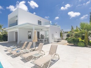 A modern two-story house with a covered patio area featuring lounge chairs and a table. - Greco Villa CCV4. 5-Schlafzimmer-Villa mit privatem Pool in Protaras Zentrum by Interhome (Protaras)