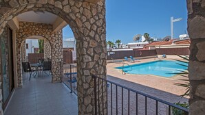 En balkon med stenvæg, ud til et poolområde med borde og stole, og udsigt til omkringliggende bygninger.