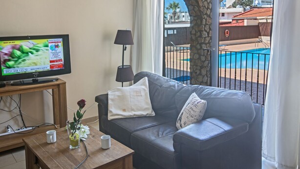 En stue med fjernsyn, sofa, et bord med blomster og udsigt til en swimmingpool og bygninger.