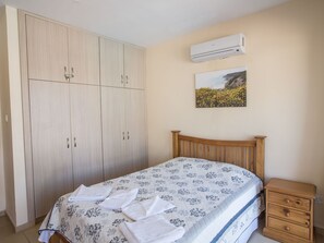 A bedroom with a bed, a wooden wardrobe, a nightstand, an air conditioning unit, and a painting on the wall. - Geräumige Wohnung mit Gemeinschaftspool by Interhome (Ayia Napa)