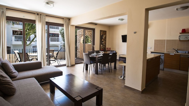 Ein modernes Wohnzimmer mit Sofa, Couchtisch und Essbereich mit Stühlen und Tisch. Es gibt eine Küchenzeile mit Schränken und Spüle. Der Raum verfügt über große Fenster und einen Blick auf ein Gebäude.