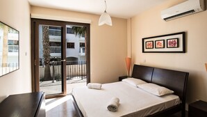 Ein Hotelzimmer mit Bett, Nachttisch, Schreibtisch, Stuhl, Fenster, Klimaanlage und einer Tür, die auf einen Balkon führt.