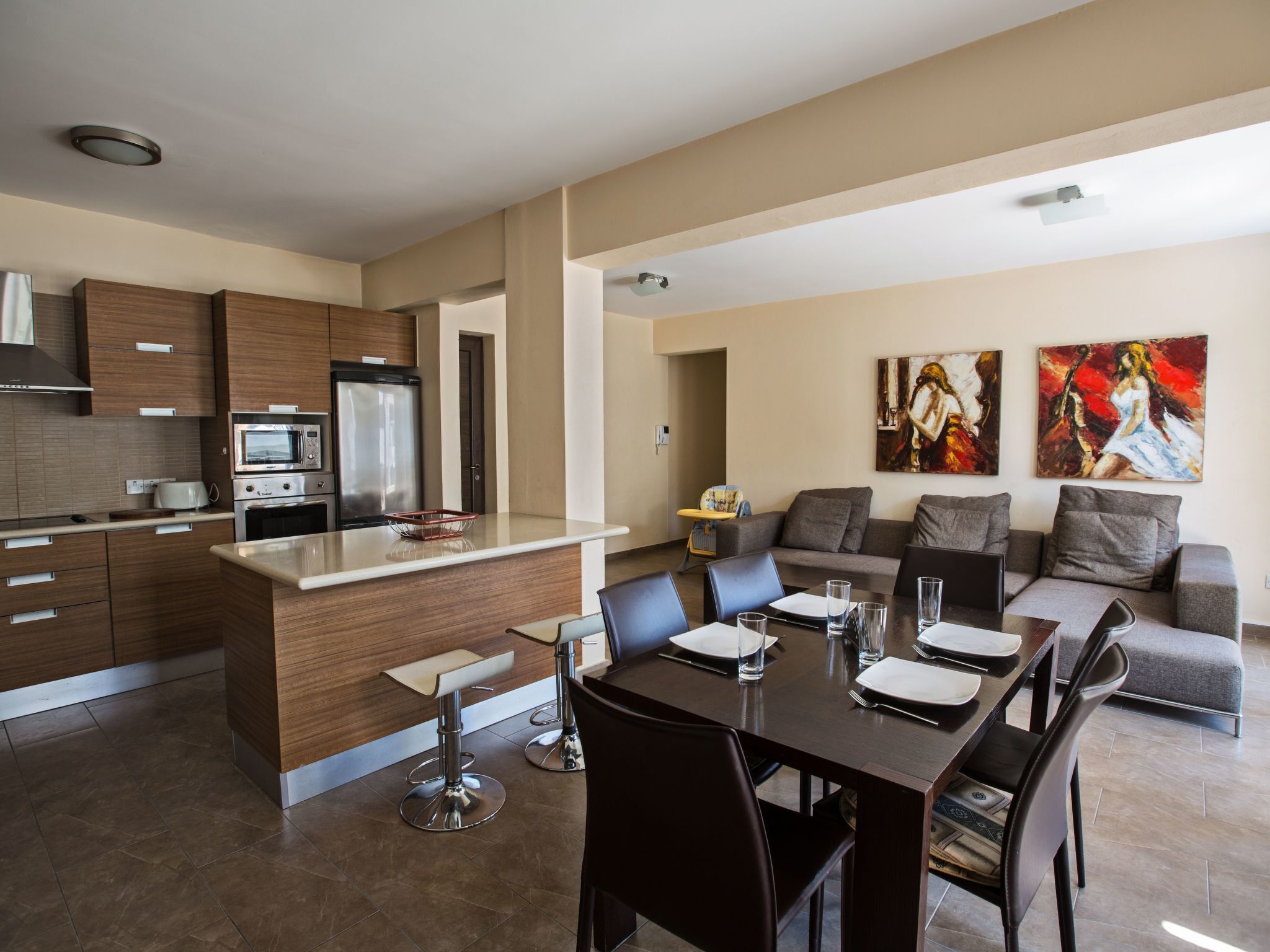 A modern kitchen with a central island, dining area with a table set for four, and a living area with a sofa and two paintings.
