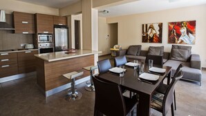 Eine moderne Küche mit einer zentralen Kochinsel, einem Essbereich mit Tisch für vier Personen und einem Wohnbereich mit Sofa und zwei Gemälden.