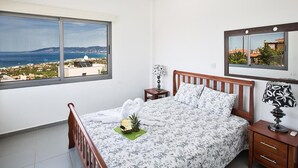 A bedroom with a bed, a nightstand, a lamp, a mirror, and a large window with a view of the sea and buildings. - Latchi Ferienvilla EV26 by Interhome (Agios Georgios Pegeias)