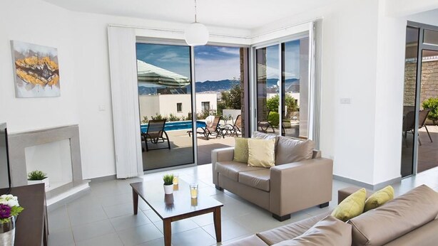 A modern living room with a sofa, a coffee table, and a fireplace. There is a large sliding glass door leading to an outdoor patio with a pool and lounge chairs. - Latchi Ferienvilla EV26 by Interhome (Agios Georgios Pegeias)