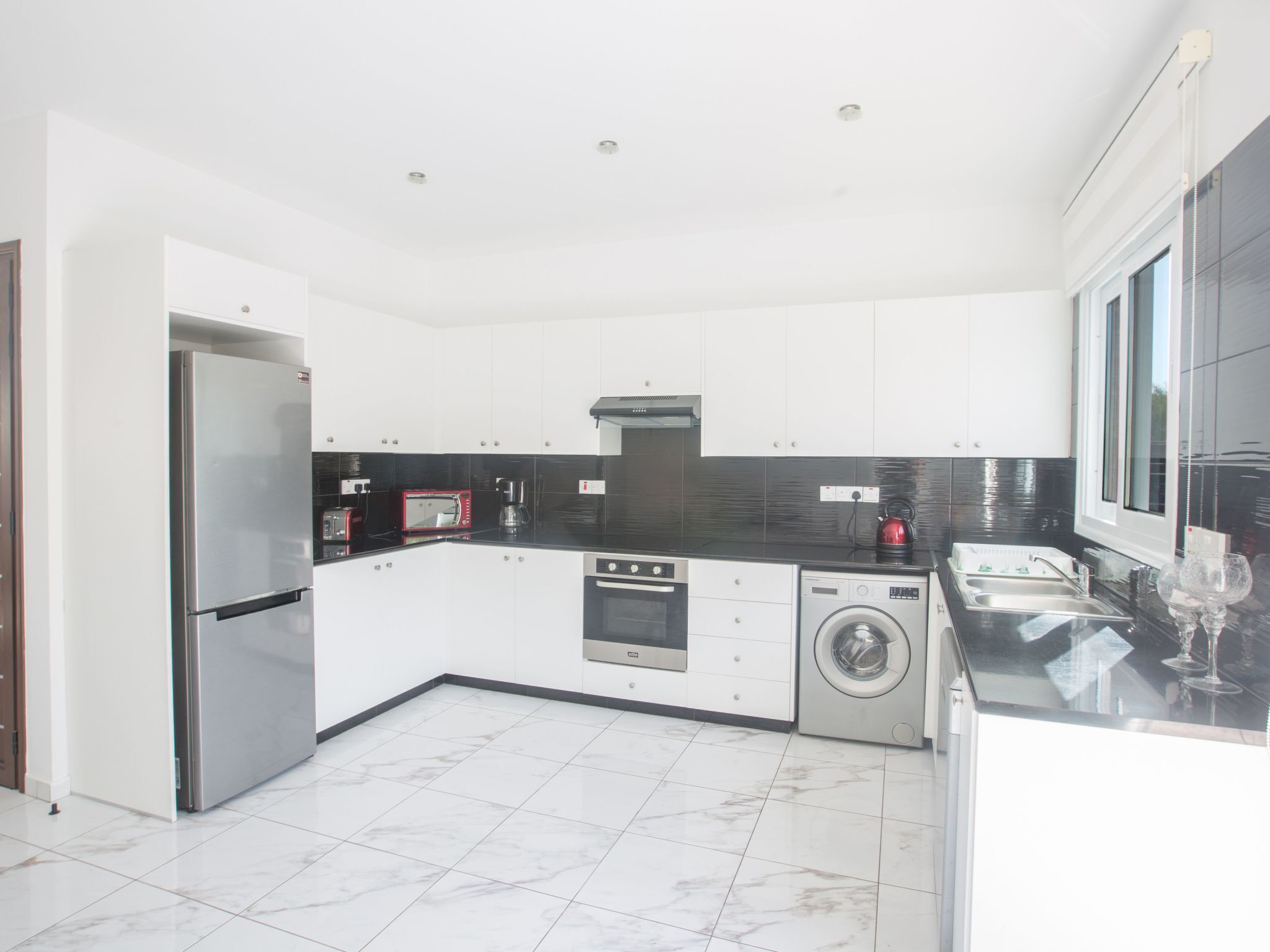 A modern kitchen with white cabinets, a stainless steel refrigerator, a built-in oven, a washing machine, and a coffee maker.