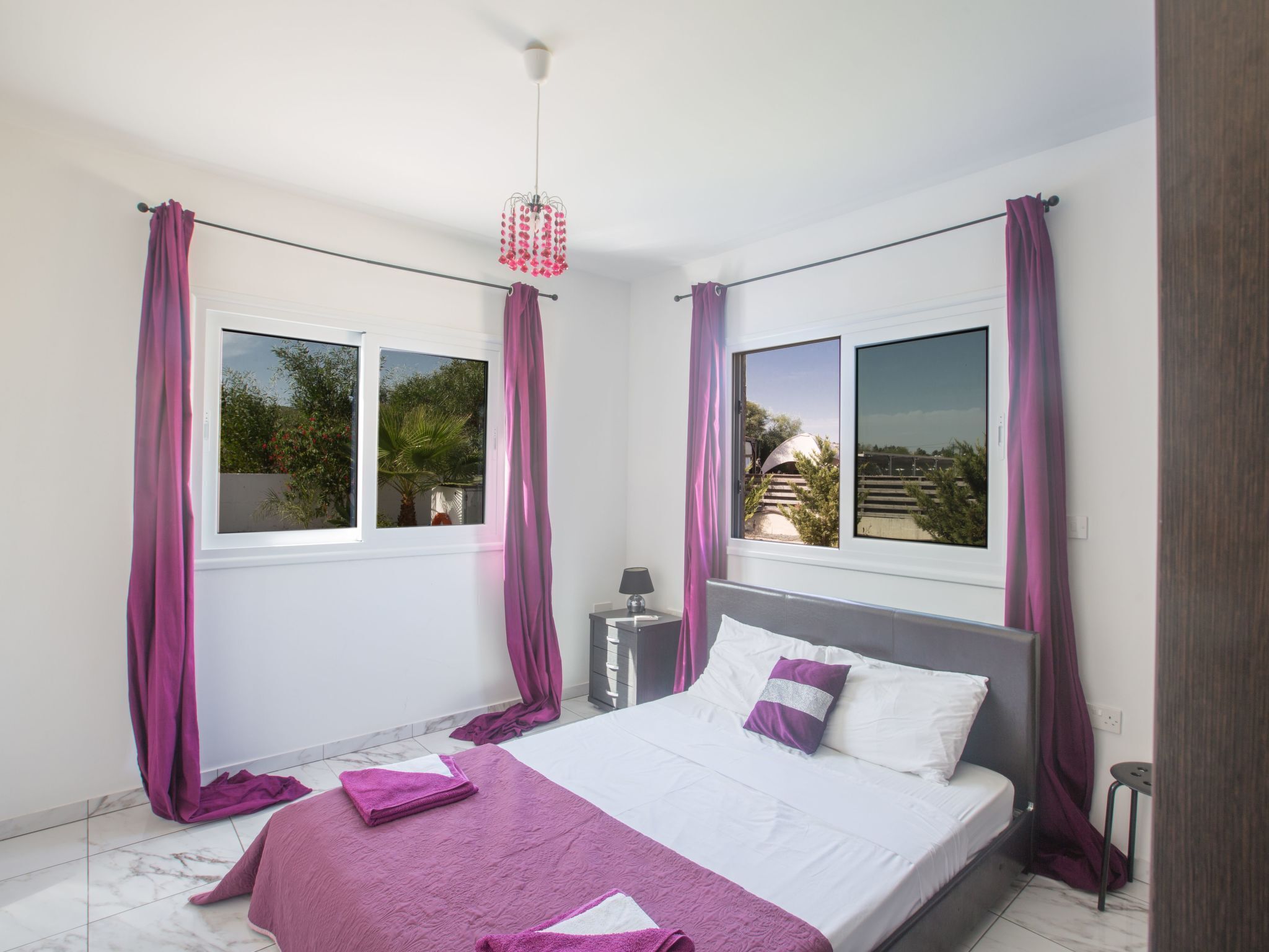 A bedroom with a bed, two windows, a nightstand, and a hanging light fixture.