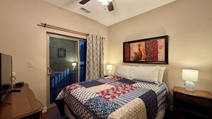Een hotelkamer met een bed, nachtkastje, bureau, raam met gordijnen, plafondventilator en een ingelijste foto aan de muur.