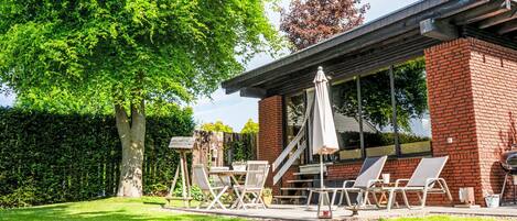 A modern outdoor patio with a brick wall, a large tree, and a covered seating area with chairs and a table.