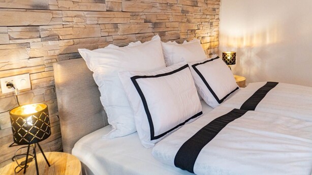 Un lit bien fait, avec une literie blanche et des bordures noires, un mur de pierre derrière, et une petite table avec une lampe placée à côté du lit.