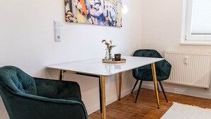 Un coin repas avec une table blanche, deux chaises vert foncé et un tableau abstrait aux couleurs vives accroché au mur.