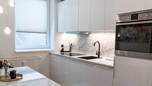 Une cuisine moderne avec des meubles blancs, un four en acier inoxydable, un plan de travail en marbre et une fenêtre équipée de stores.