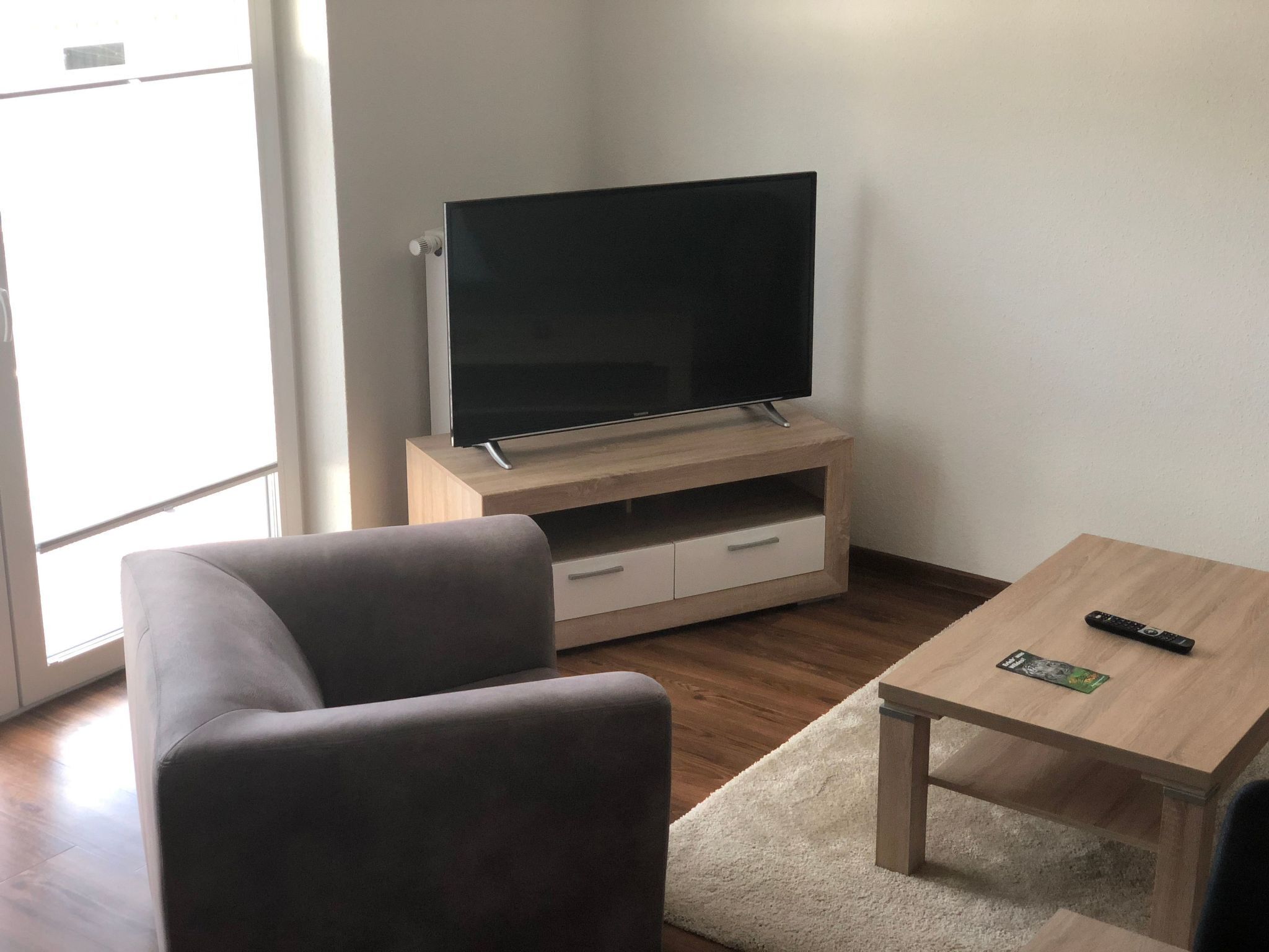 A living room with a flat-screen TV on a wooden stand, a grey sofa, a small wooden table with a remote, and a white rug.