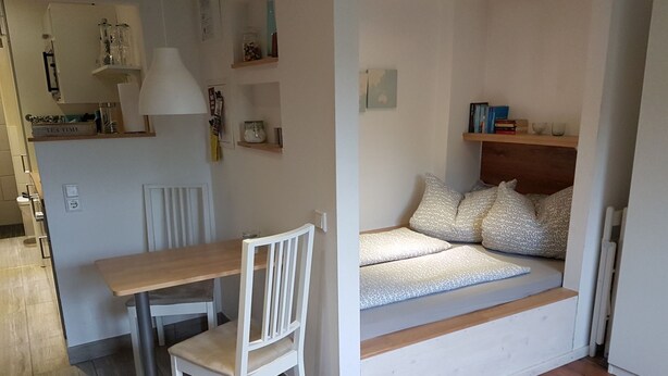 A compact living space with a wooden dining table, a white chair, a built-in bench with patterned cushions, and shelves with various items. - Apartment Oliver by Interhome (Fritzlar)