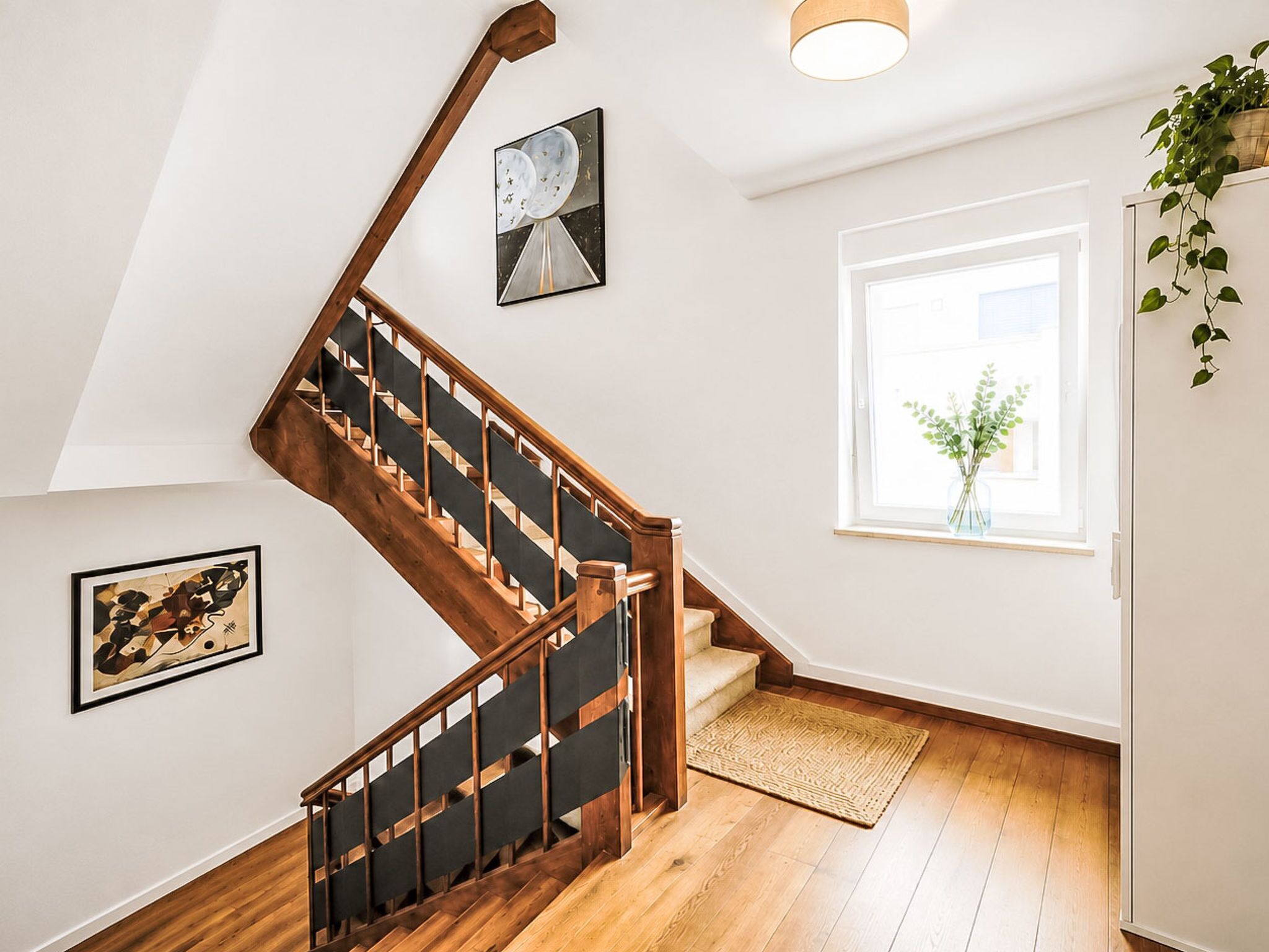 Un escalier avec des rampes en bois et une main courante noire, menant à un palier où est accrochée une œuvre d’art encadrée, et une fenêtre donnant sur un paysage verdoyant.