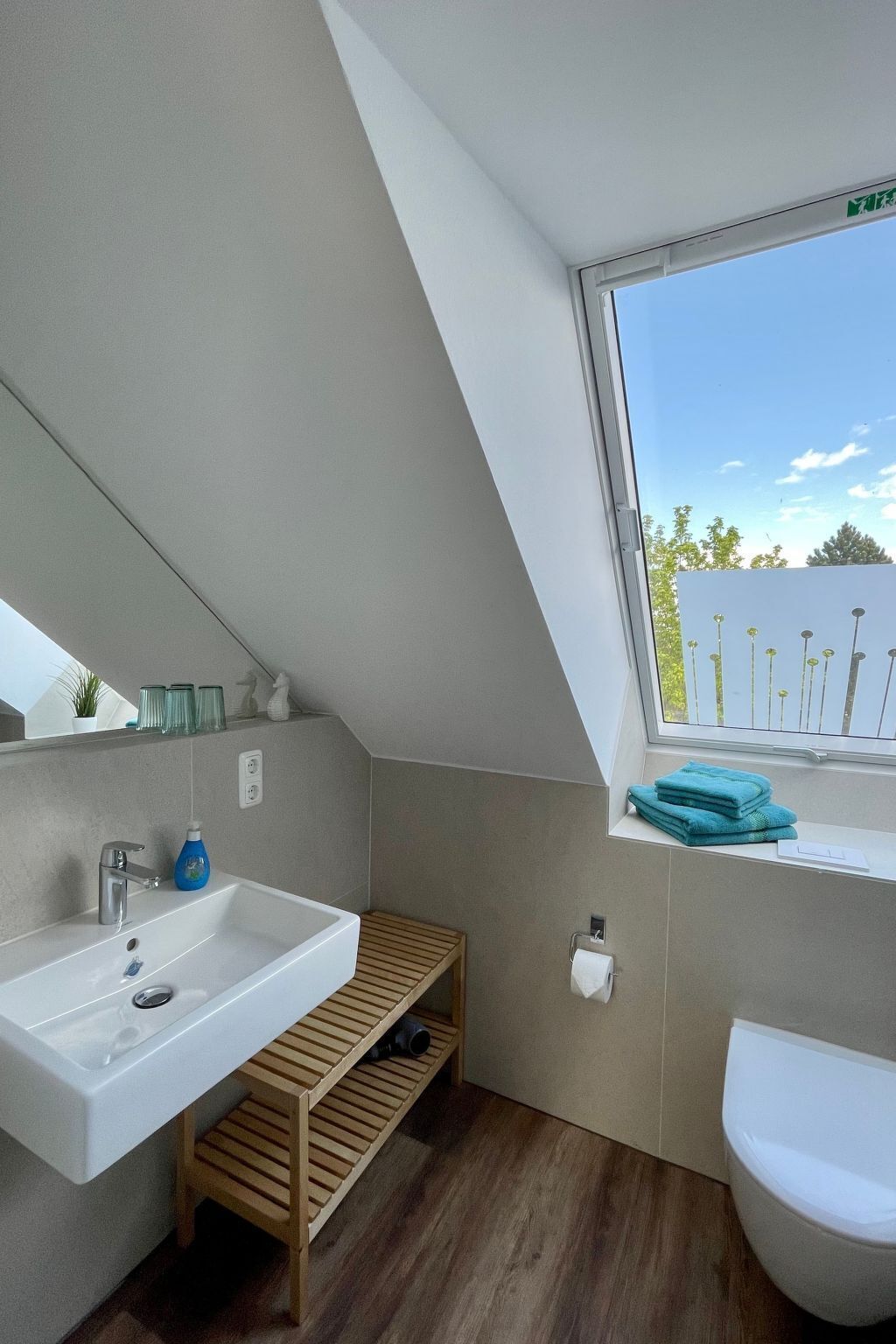 Une salle de bain moderne avec un lavabo blanc, une étagère en bois et une fenêtre donnant sur les arbres.