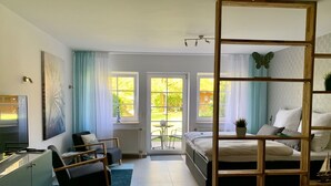 Ett modernt sovrum med en säng, ett skrivbord och en stol. Det finns två fönster med gardiner, utsikt över trädgården och en avskärmande vägg i trä.