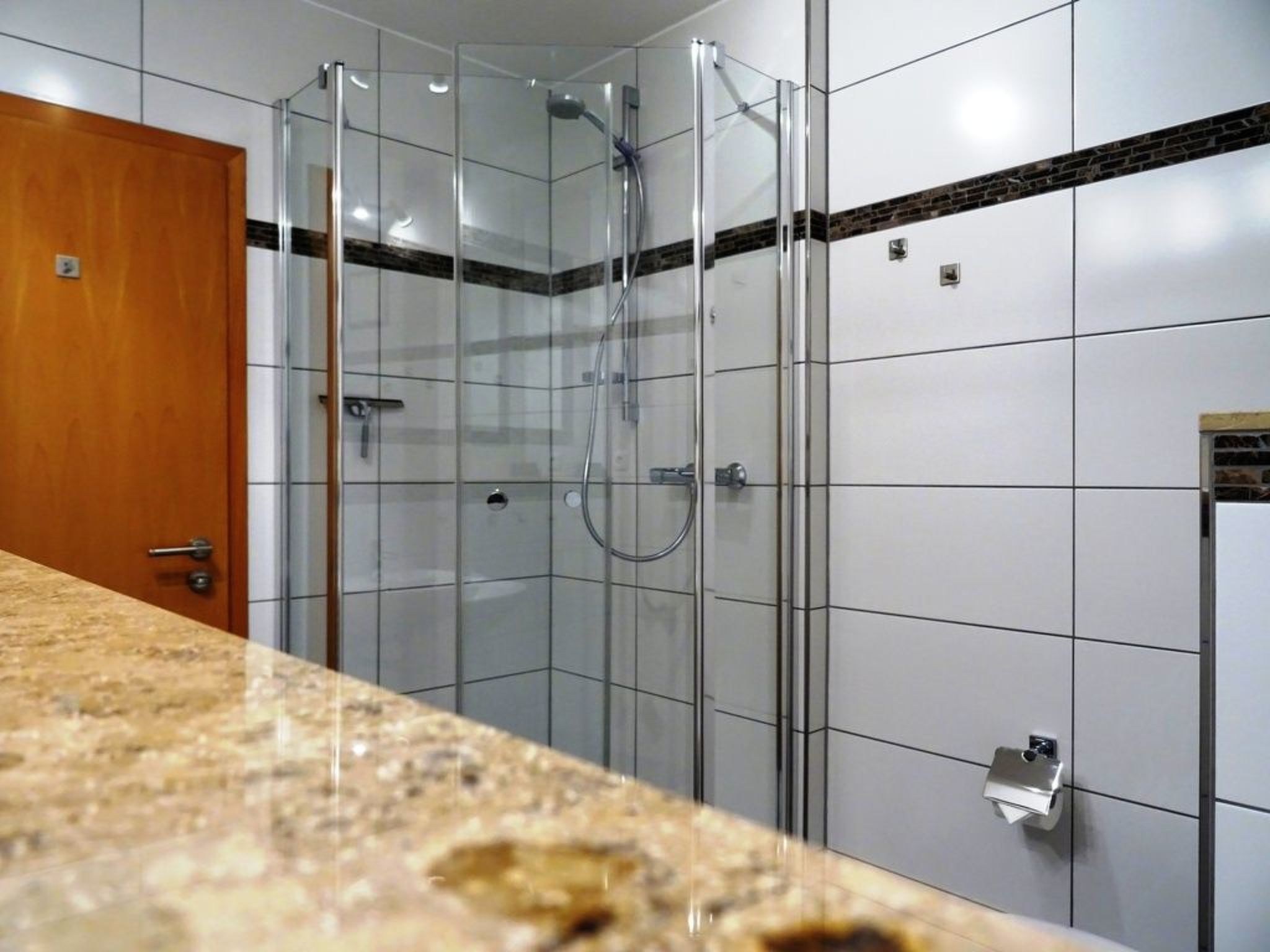 Une salle de bain dotée d’une douche à parois de verre, de murs recouverts de carreaux blancs, d’une porte en bois et d’un comptoir en granit.