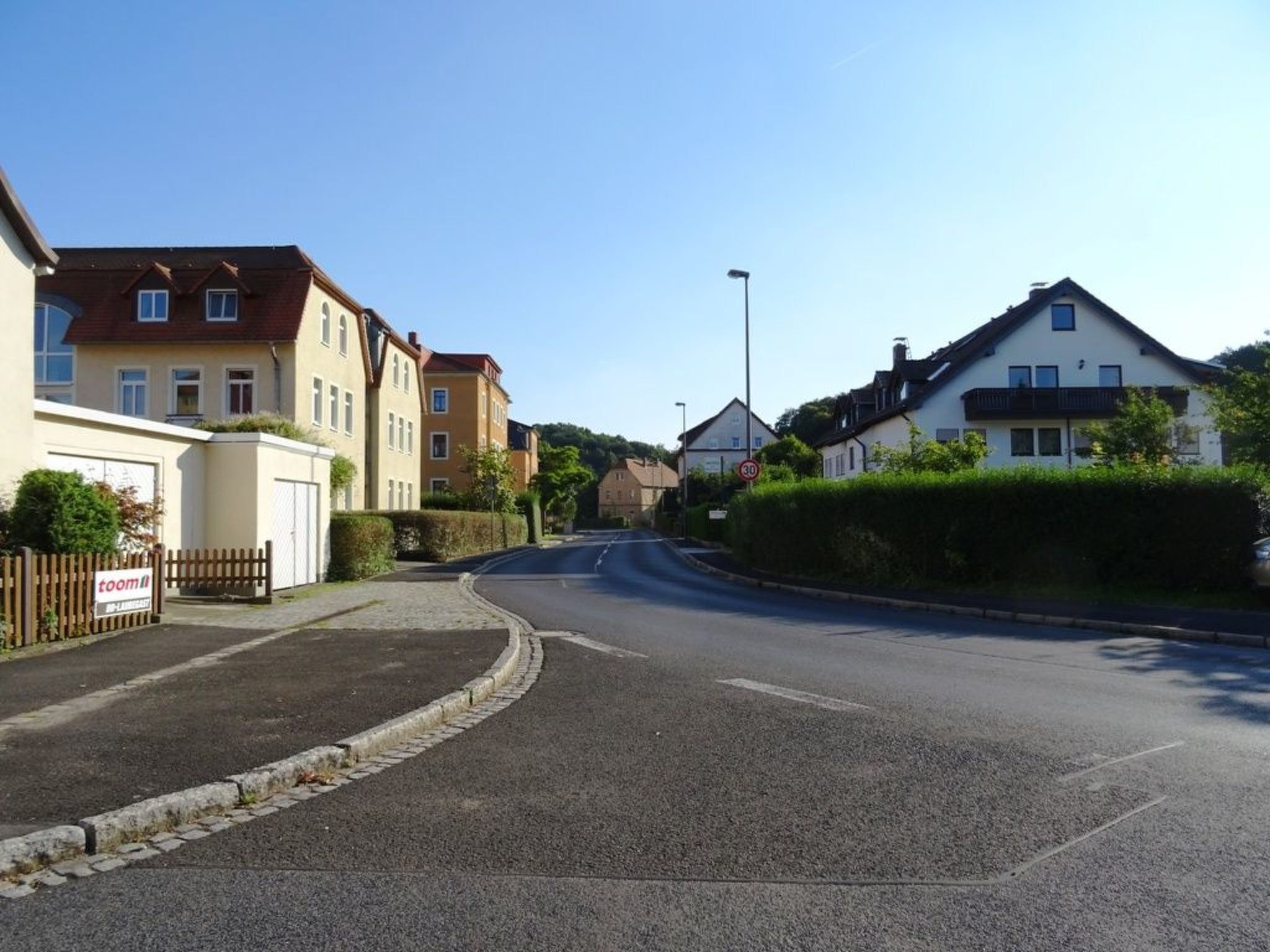 Une rue résidentielle bordée de maisons des deux côtés, un ciel dégagé et un panneau indiquant une limitation de vitesse à 30.